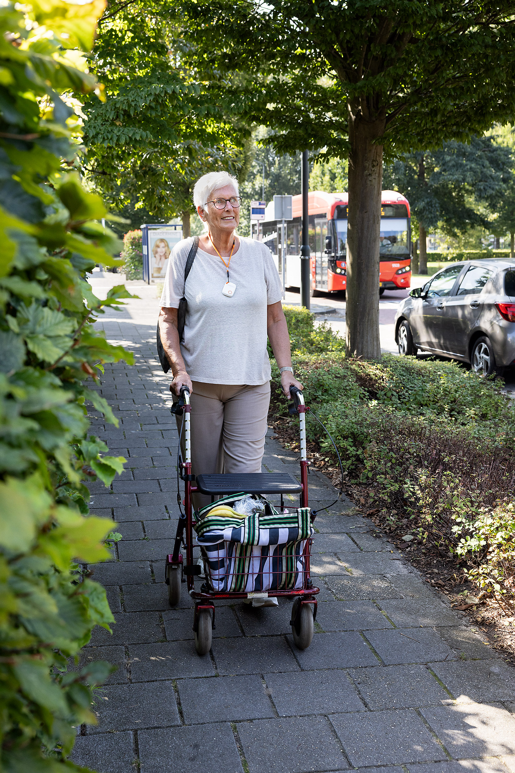 Senior dame met persoonsalarm Navi loopt met rollator op de stoep. Op de achtergrond is een bus te zien.